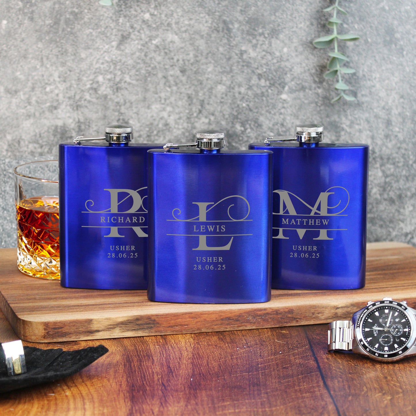 Three blue flasks with engraved names on a wooden surface with a glass of whiskey.