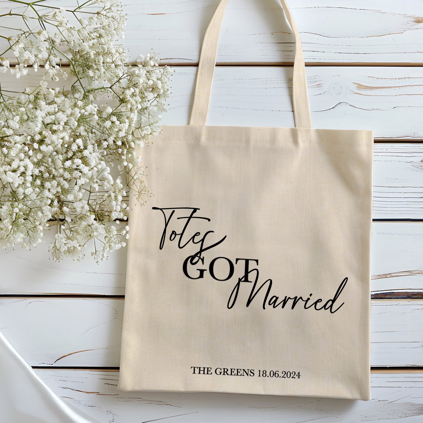 Beige tote bag with 'Totes Got Married' text on a wooden surface with white flowers.