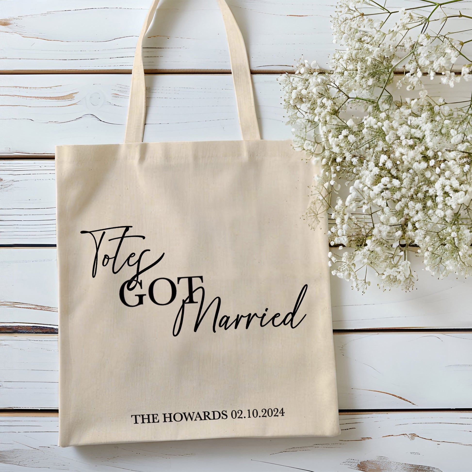 Beige tote bag with 'Totes Got Married' text and personalized details on a wooden surface with white flowers.