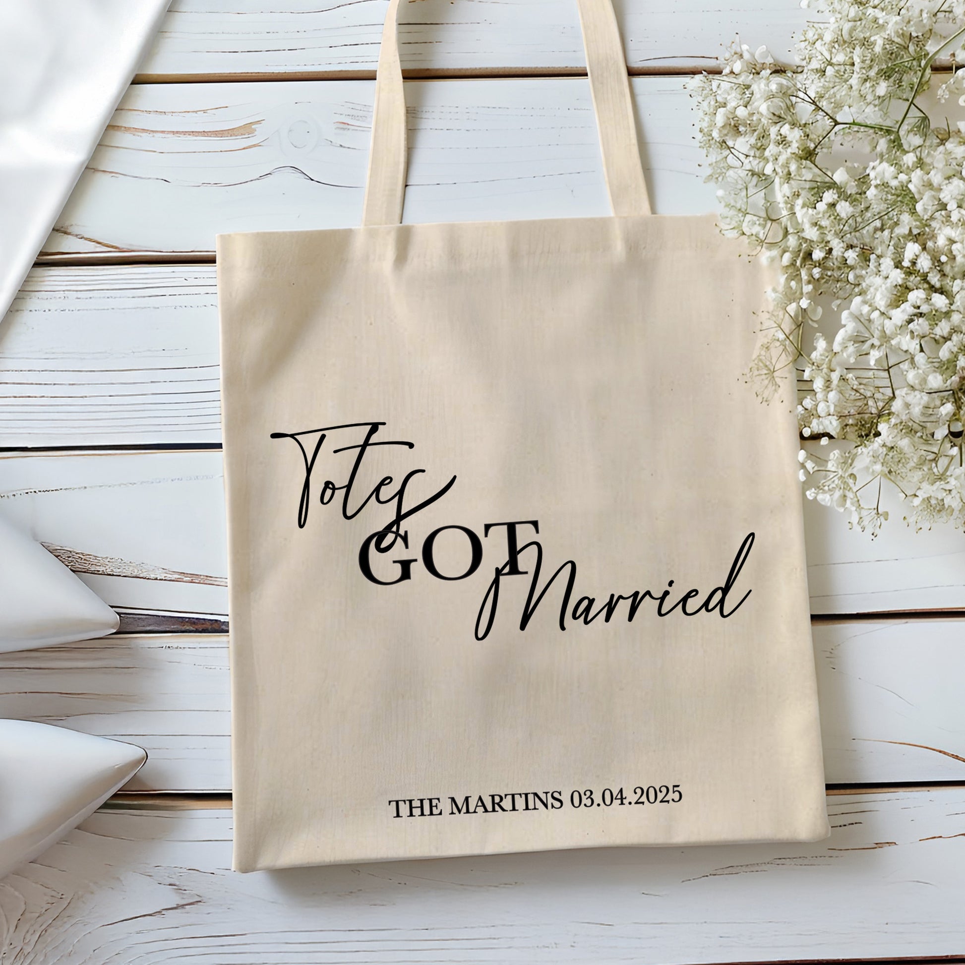 Beige tote bag with 'Totes Got Married' text on a wooden surface with white flowers.