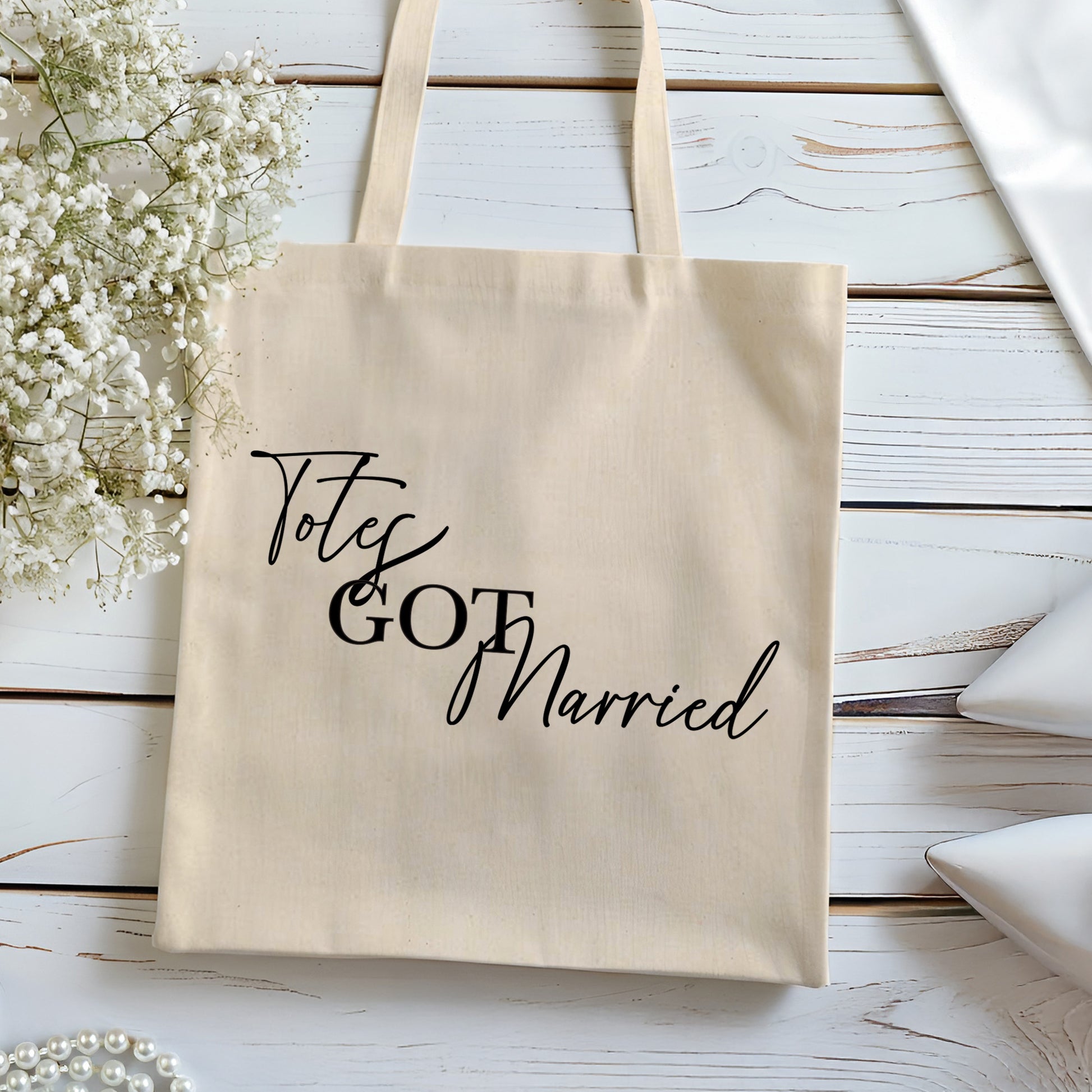Beige tote bag with 'Totes Got Married' text on a wooden surface with flowers.