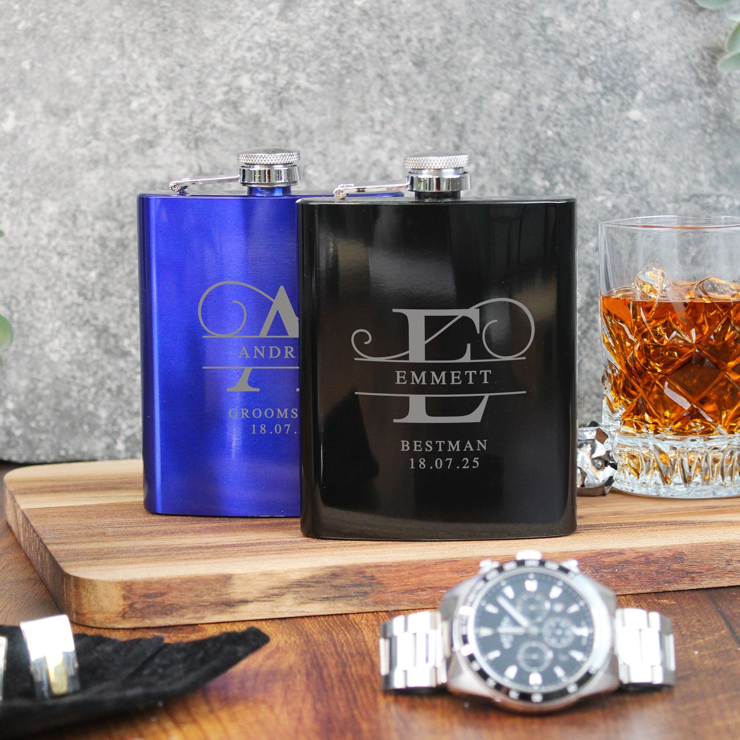 Two flasks with engraved names and a glass of whiskey on a wooden surface.