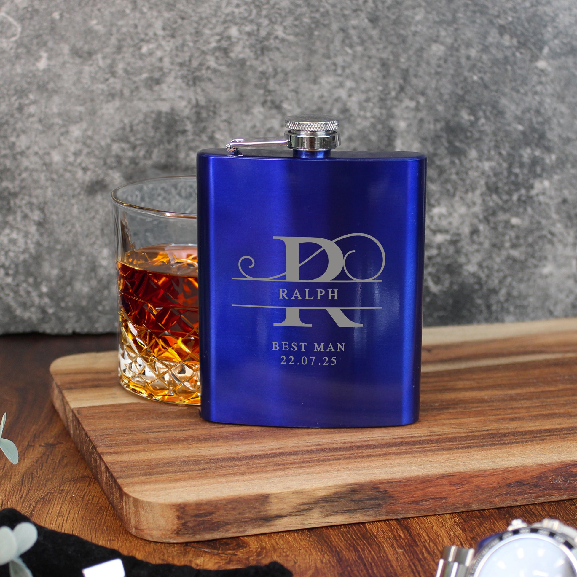 Blue flask with engraved text next to a glass of whiskey on a wooden surface.
