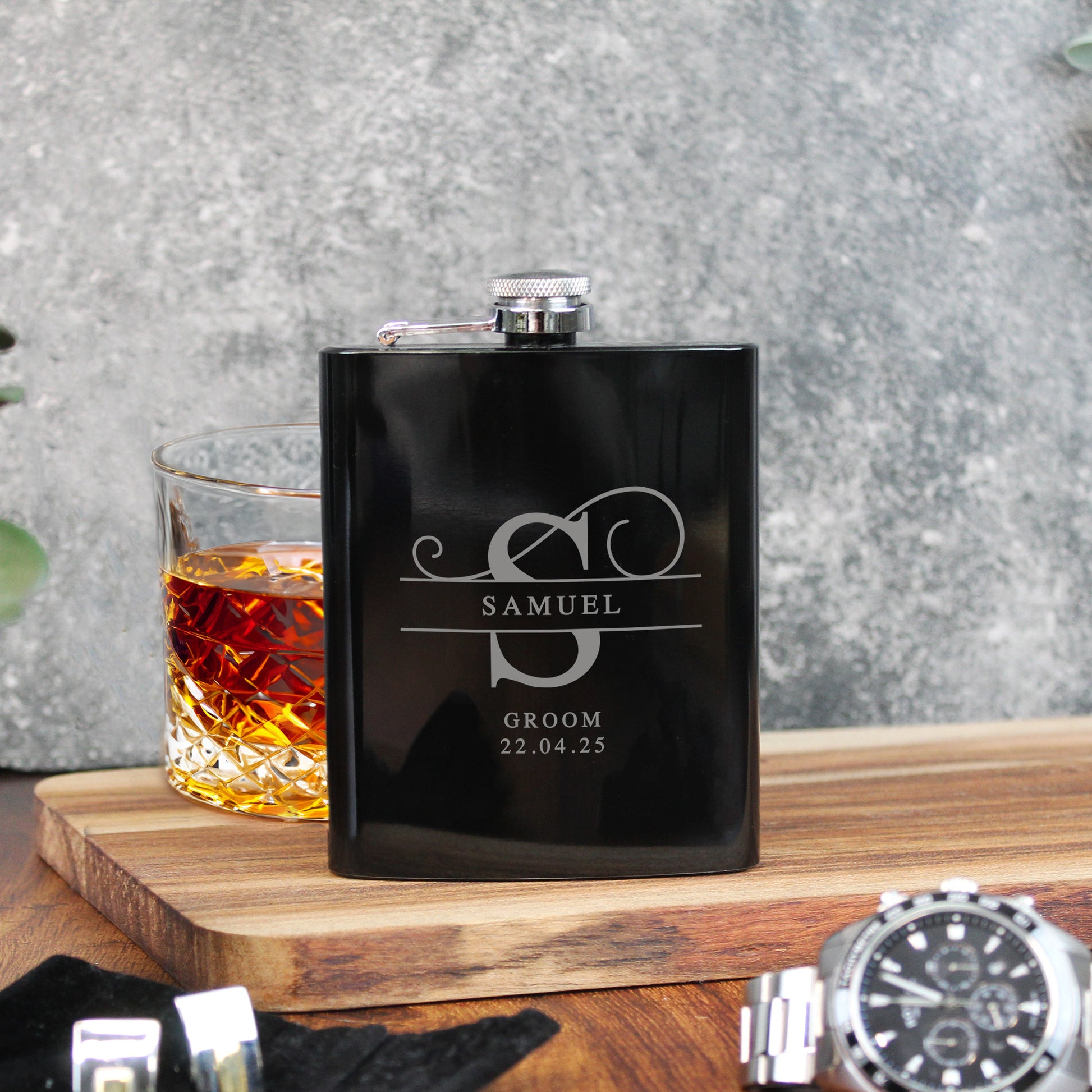 Black flask with engraved text on a wooden surface with a glass of whiskey.