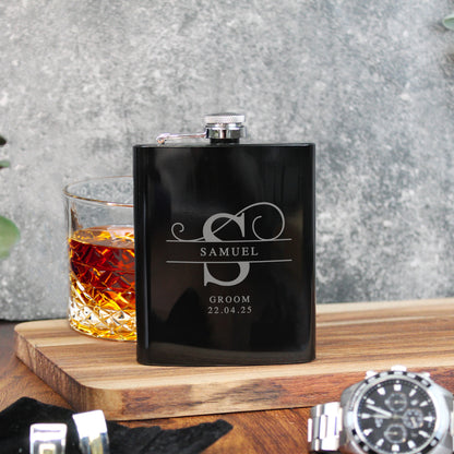 Black flask with engraved text on a wooden surface with a glass of whiskey.
