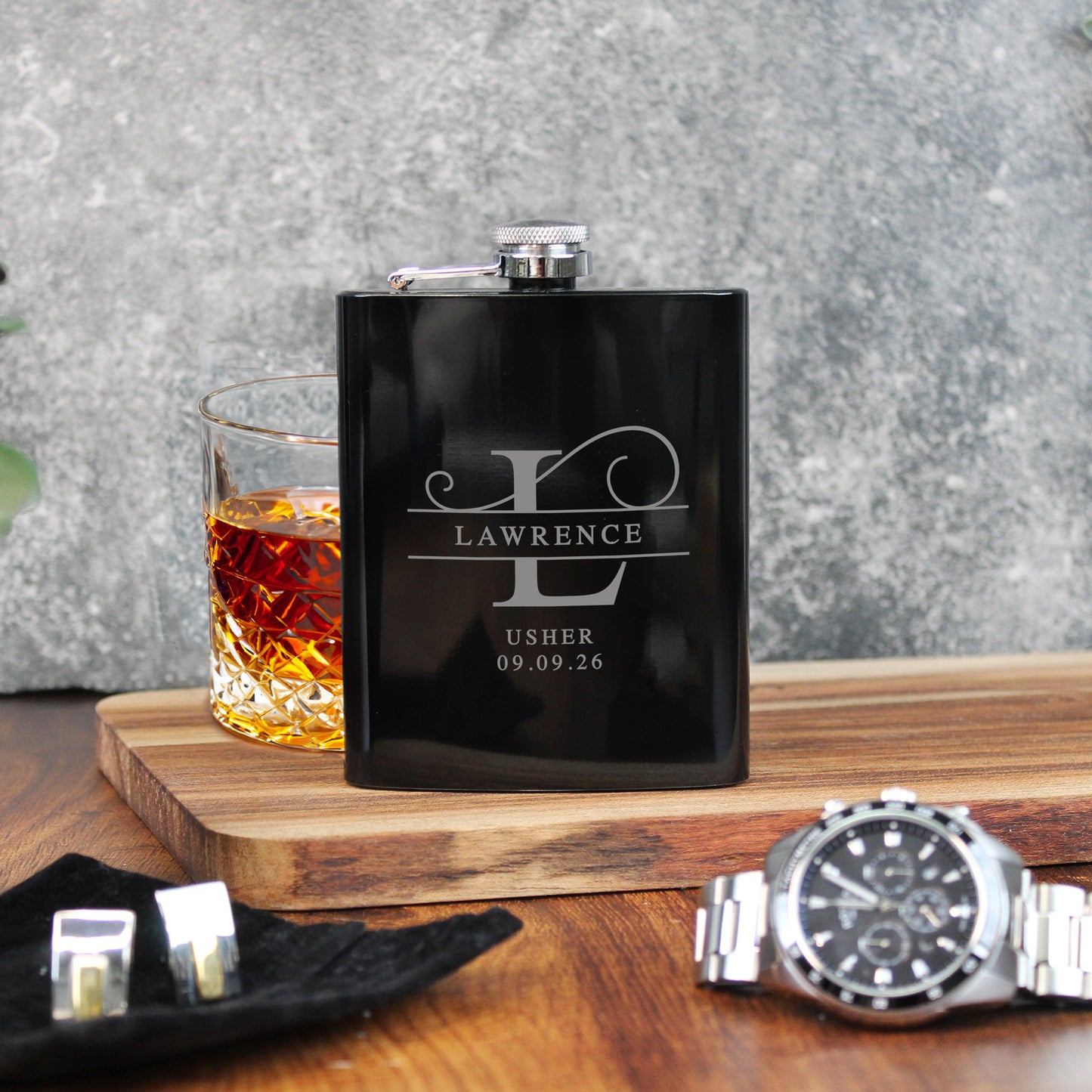 Black flask with engraved name and date, glass of whiskey, and watch on a wooden surface.