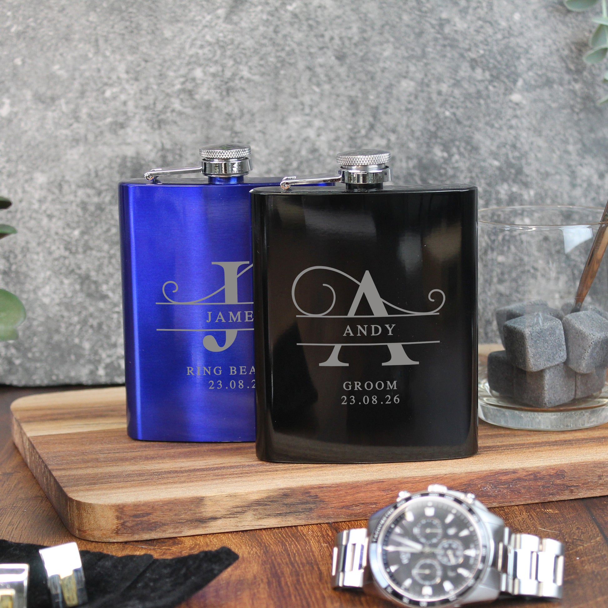 Two personalized flasks, one blue and one black, on a wooden surface with a watch in the foreground.