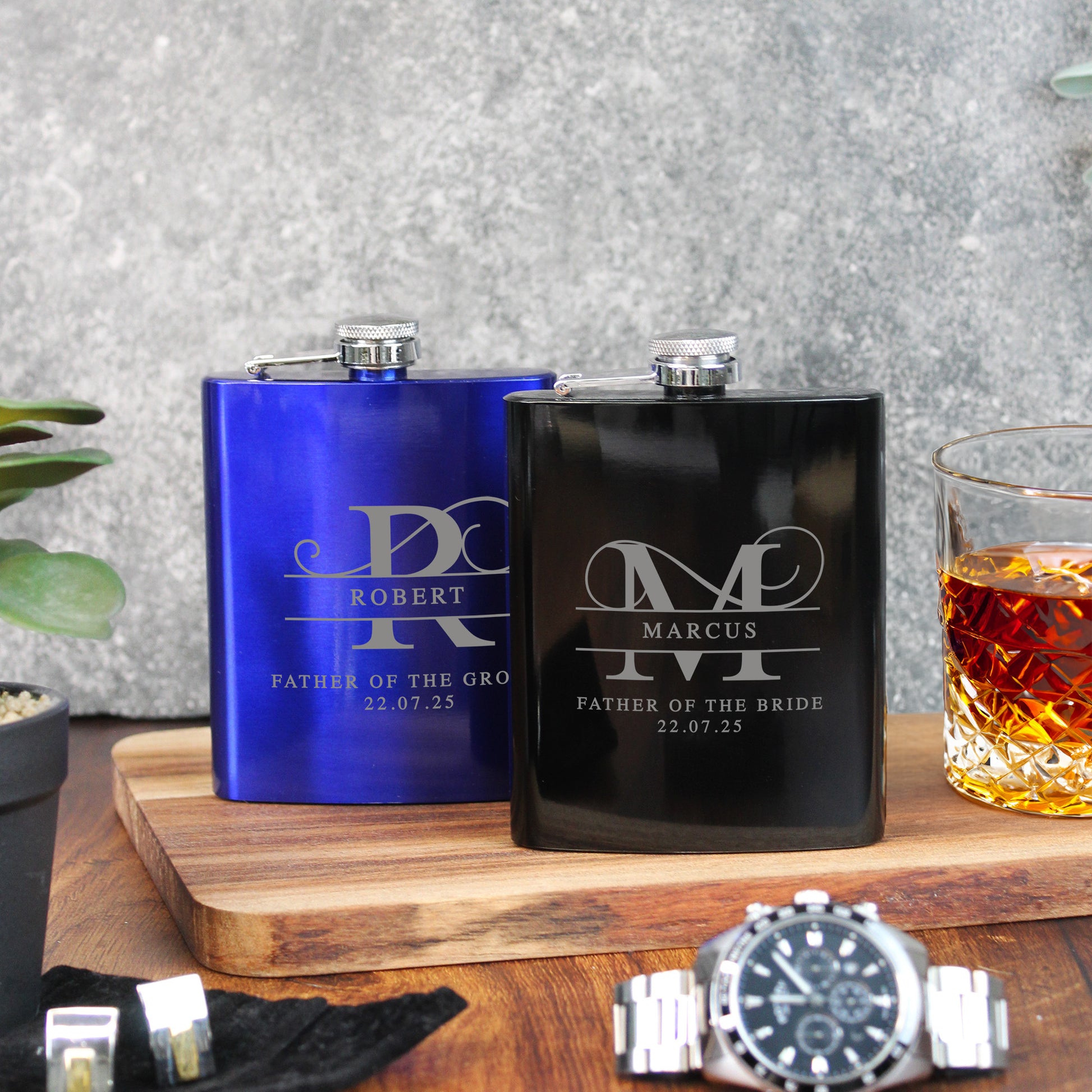 Two personalized flasks, one blue and one black, on a wooden surface with a glass of whiskey.