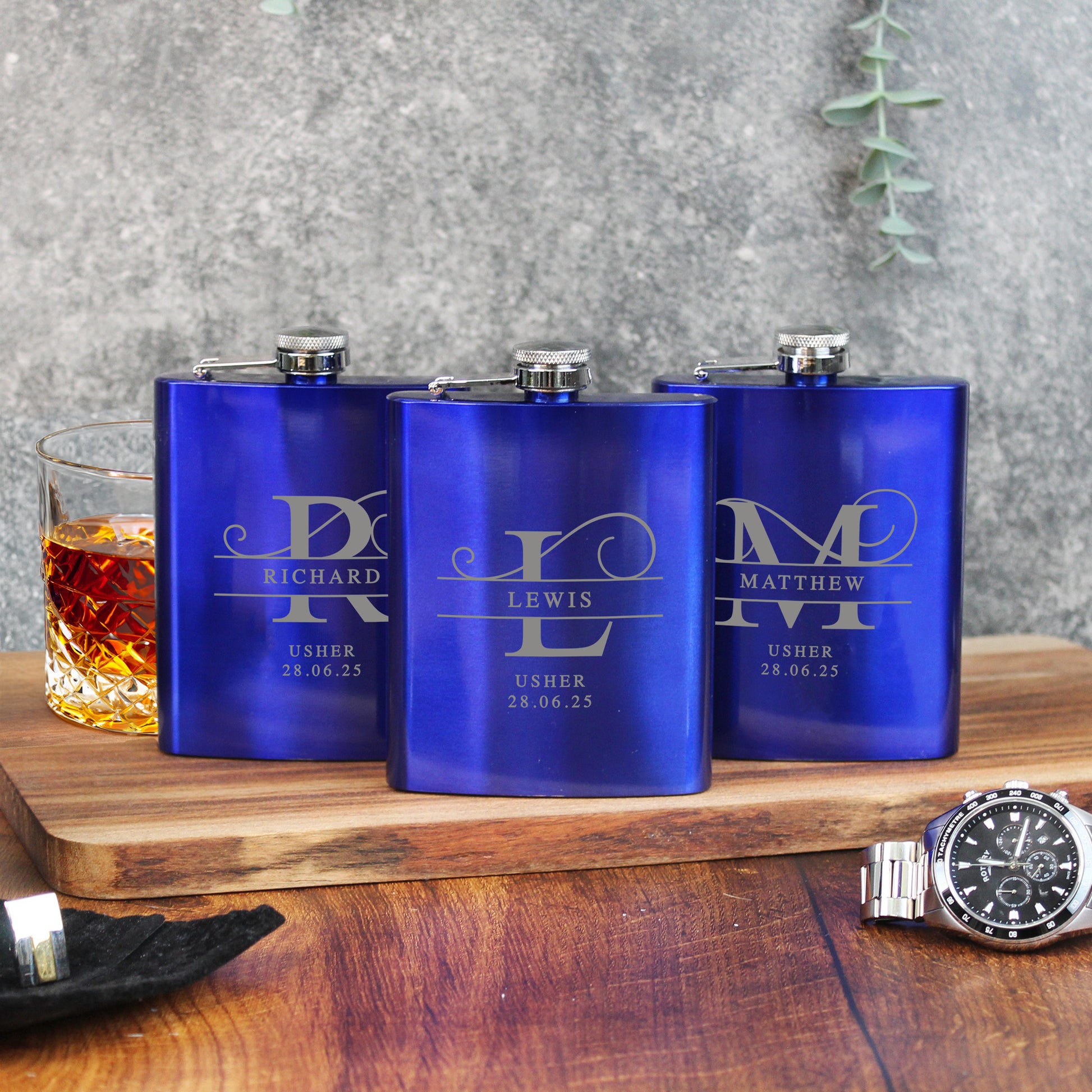 Three blue flasks with engraved names on a wooden surface with a glass of whiskey.