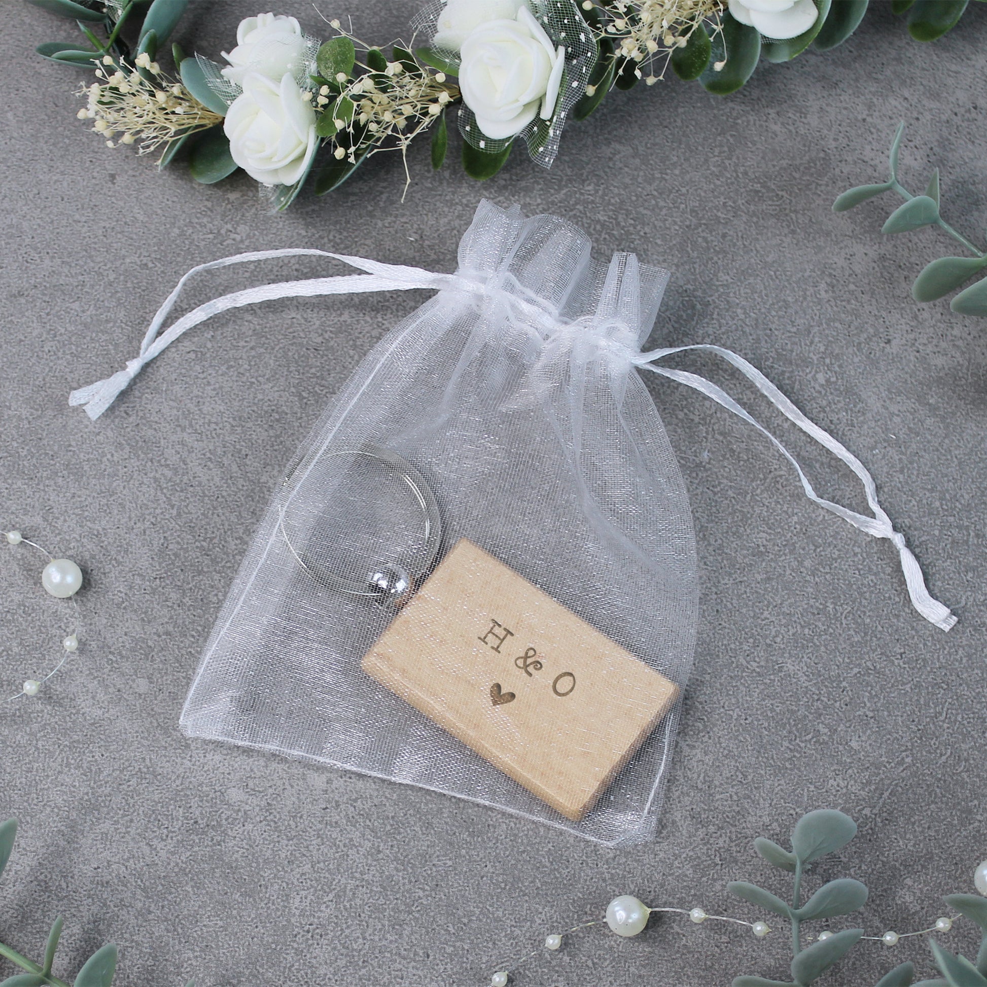 Wooden block with 'H & O' engraving in a clear drawstring bag on a gray surface with floral decorations.