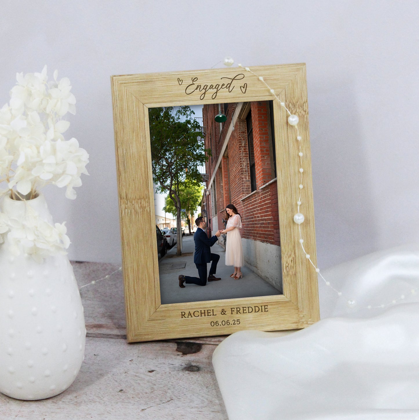 Wooden photo frame with engraved text displaying a couple getting engaged, placed on a surface with decorative elements.