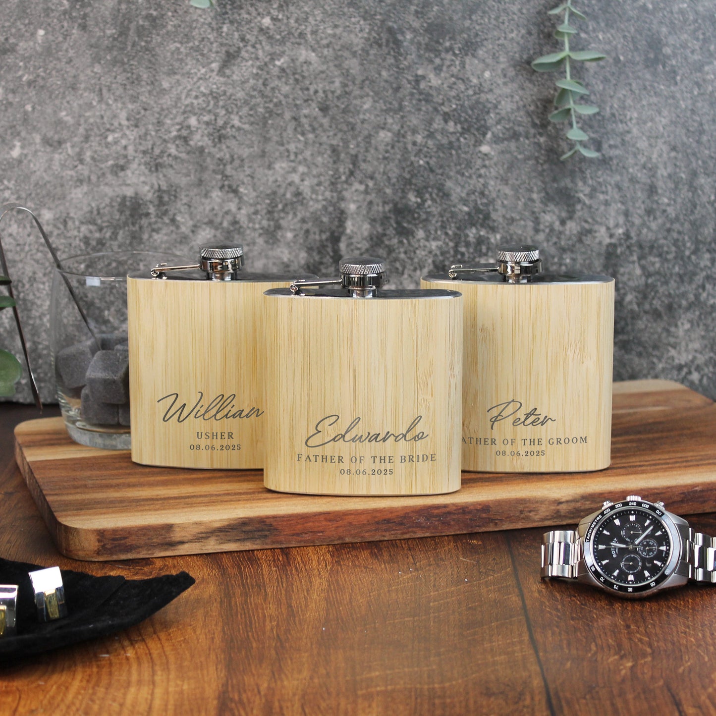 Three wooden flasks with engraved names on a wooden surface with a watch and glasses in the background.