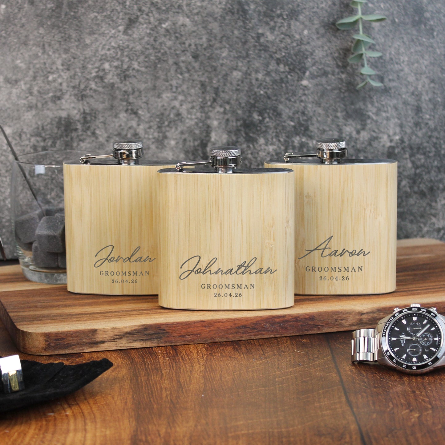 Three wooden flasks with engraved names on a wooden surface with a watch in the foreground.