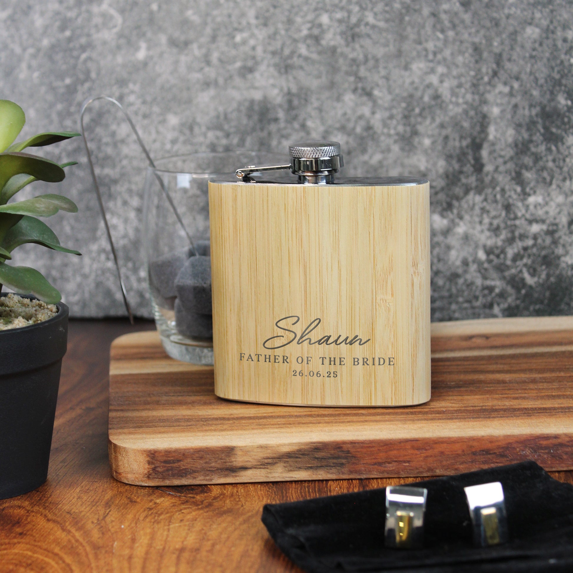 Wooden flask with engraved text on a wooden surface with a plant and glass in the background.